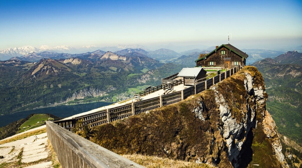 Schafberg cog rail way (c) Schafber Wooden mountain lodge on a cliff edge overlooking a scenic valley and distant mountains under a clear sky. (Enlarged view)