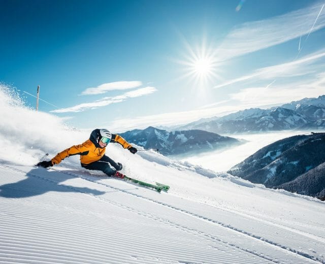 Skier in yellow jacket carving down a groomed snowy piste in Zell am See, sunlit mountains in the background.