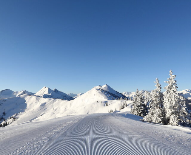 A freshly groomed ski piste leads to snow-covered mountains under a clear blue sky.