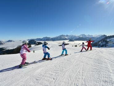 Four children in ski kit follow an instructor down Saalachtal’s snowy mountain under a clear blue sky.