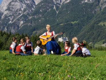 A woman plays guitar on grass, Sound of Music style, surrounded by children in Austrian traditional clothes, mountains behind.