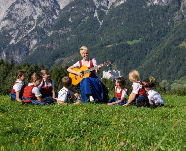 The Sound of Music A woman plays guitar on grass, Sound of Music style, surrounded by children in Austrian traditional clothes, mountains behind.