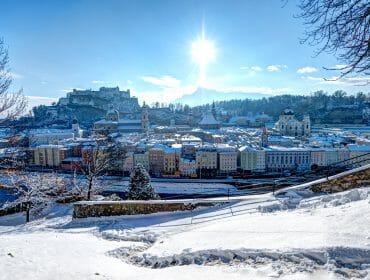 Snow-covered Salzburg cityscape with the sun shining above, castle and Salzburg’s surrounding mountains.