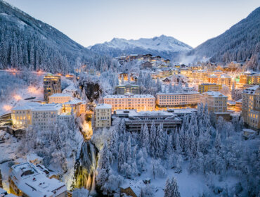 Snow-covered Gastein town nestled in alpine mountains with lit buildings and a waterfall at dusk.