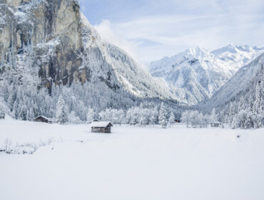 Snow-covered Gastein valley with scattered cabins, pine forests, and tall, rocky mountains under a blue sky.
