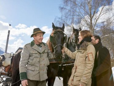 Two people pat a horse by a carriage and driver in snowy Stuhlfelden with trees and mountains.
