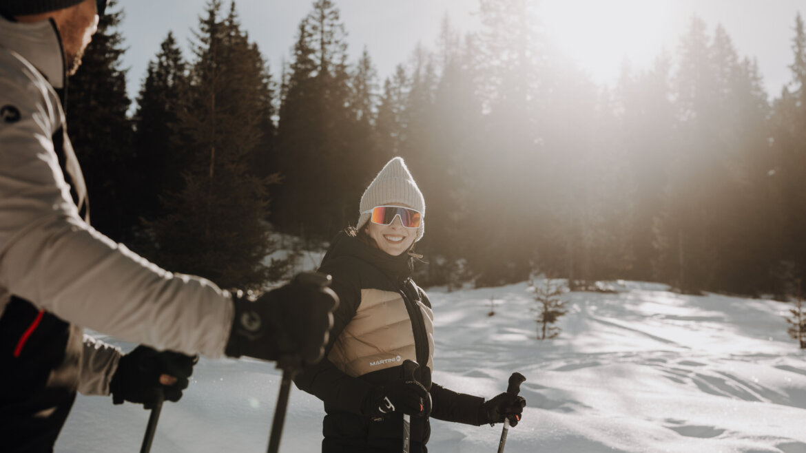 © Tourismusverband Bischofshofen | two minds Studio Two people in winter gear ski through a snowy forest, sunlight shining behind tall evergreen trees. (Enlarged view)
