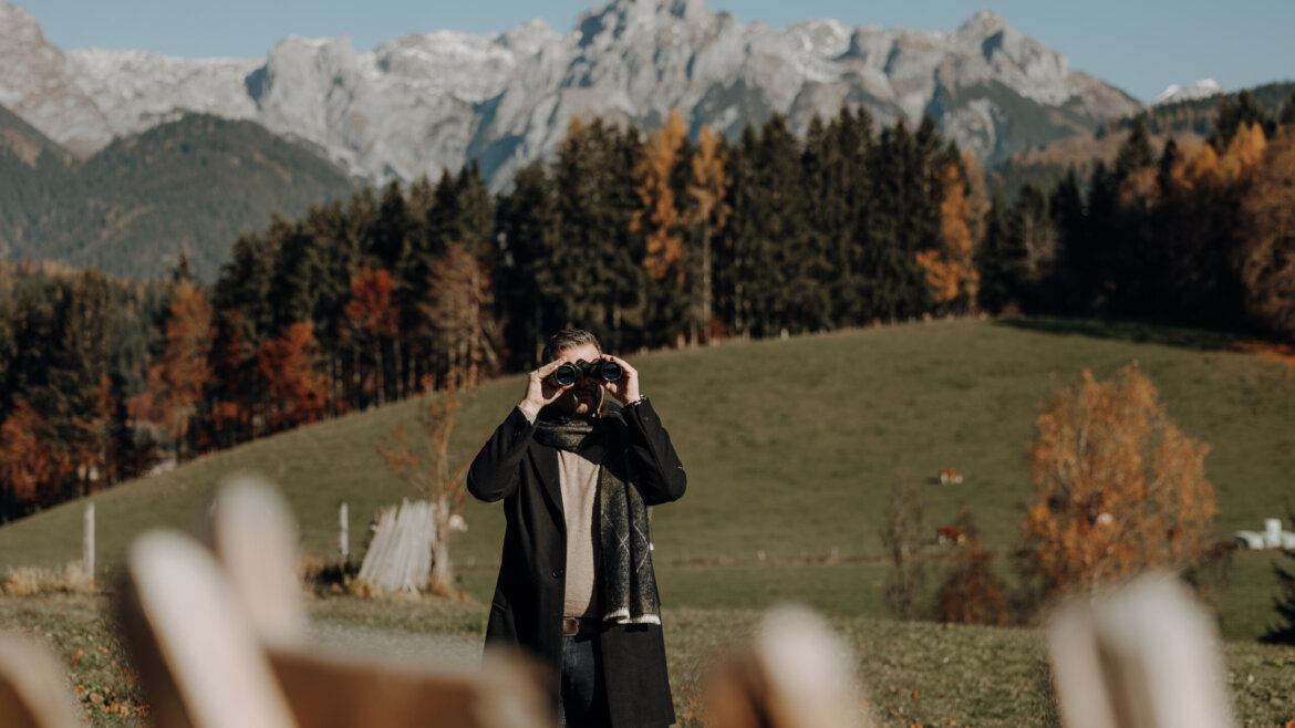 © Tourismusverband Bischofshofen | two minds Studio Person in a coat and scarf uses binoculars in a grassy field with mountains and trees in the background. (Enlarged view)