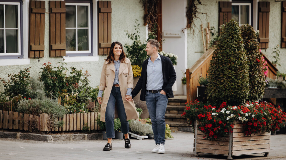 A smiling couple walk hand in hand on a quaint street lined with flowers. (Enlarged view)