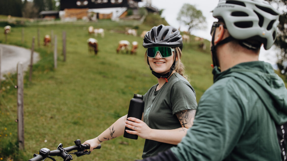 Two cyclists in helmets pause on a rural path, one smiling and holding a water bottle, cows grazing nearby. (Enlarged view)