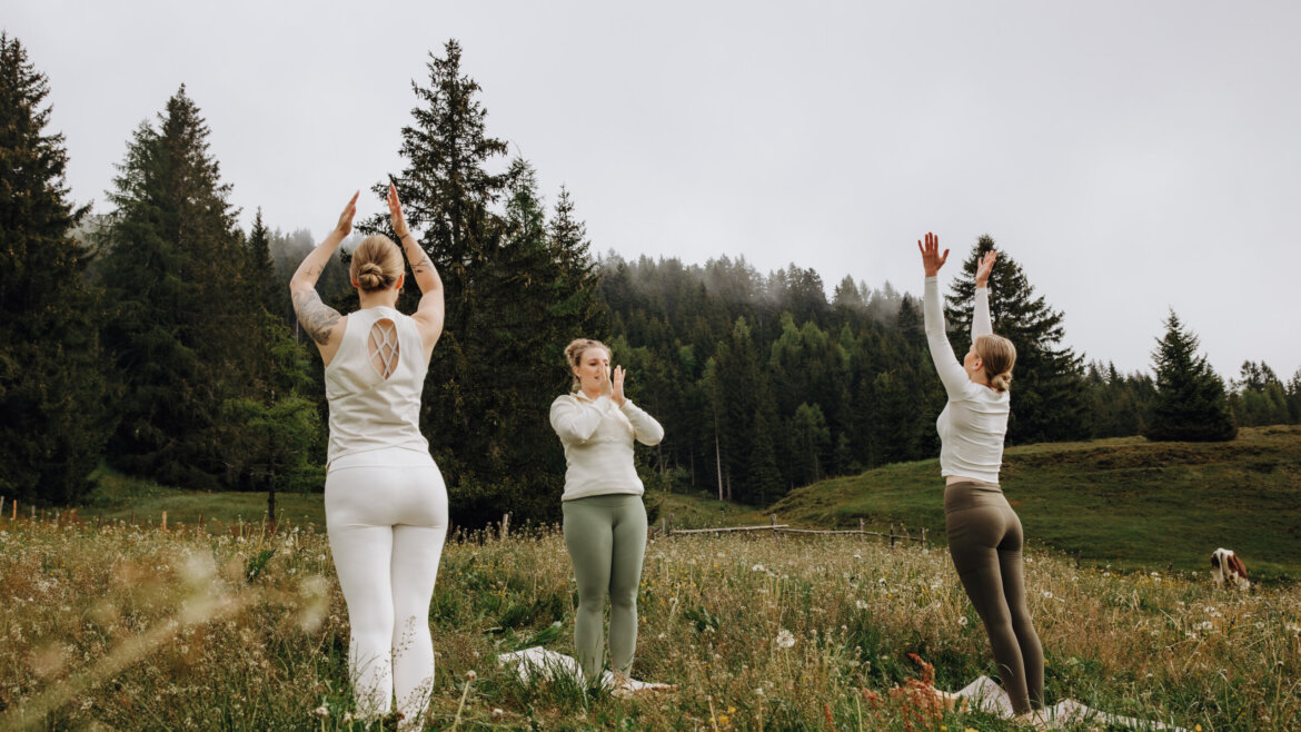 Three women practise yoga outdoors on a grassy field surrounded by trees and mountains under a cloudy sky. (Enlarged view)