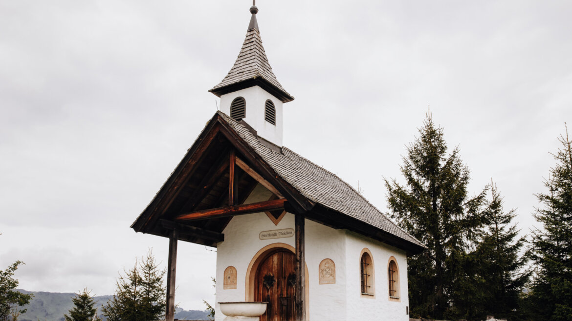 The Meislsteinkapelle, a small white chapel with a wooden roof and spire, surrounded by trees and a wooden fence under a cloudy sky. (Enlarged view)