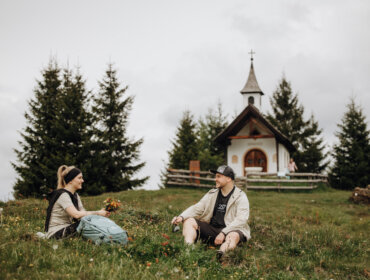 Two people sit on grass, chatting with the chapel "Meislsteinkapelle" and pine trees in the background.