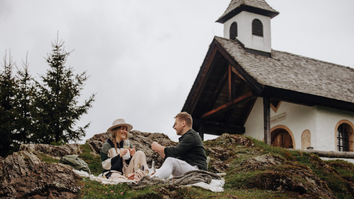 A couple enjoys a picnic on a grassy hill near a small chapel, the Meislsteinkapelle, with a spire on a cloudy day. (Enlarged view)