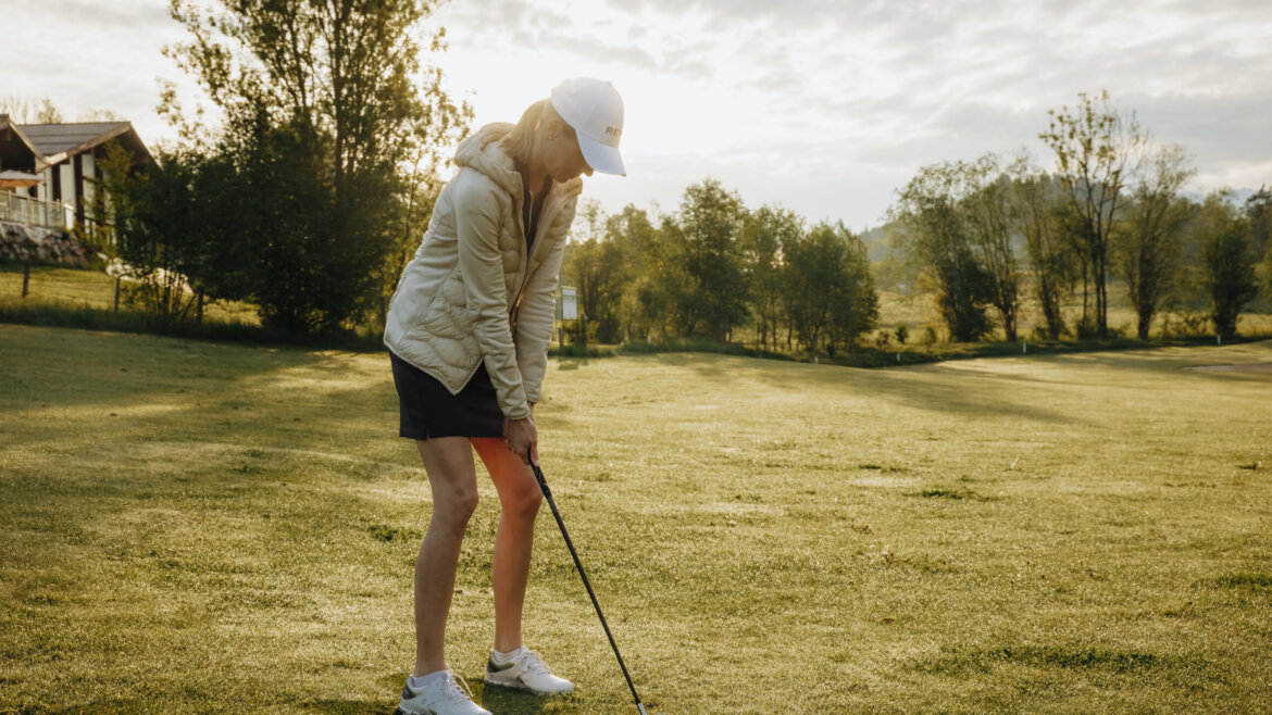 Woman in a cap and jacket preparing to hit a golf ball on a grassy course at sunrise or sunset. (Enlarged view)
