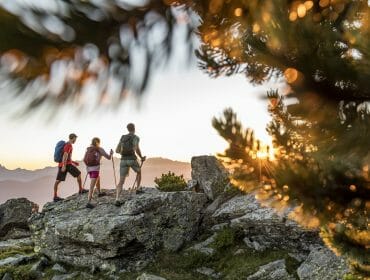 Three hikers with rucksacks walk on rocky terrain at sunset near Flachau, framed by trees and distant mountains.