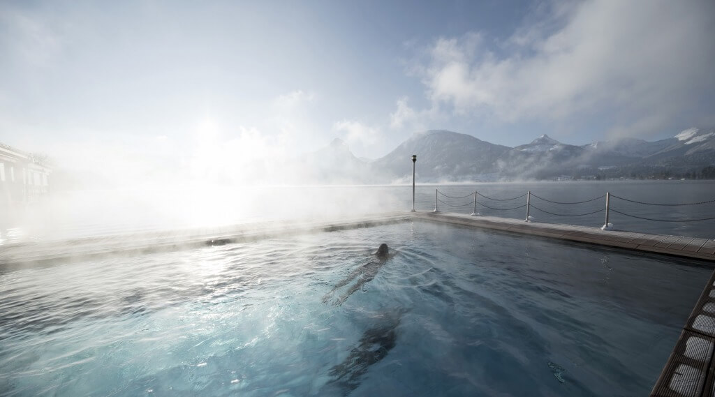 Weißes Rössl outdoor schwimmingpool (c) oberösterreichtourismusgmbh davidlugmayr A person swims in a steaming outdoor pool overlooking a misty lake and snow-capped mountains. (Enlarged view)