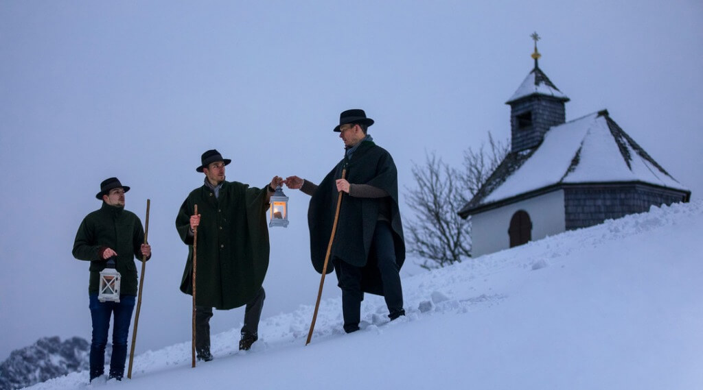 Winter advent (c) SalzburgerLand Tourismus Three people holding lanterns and walking in the snow near a small chapel on a hillside at dusk. (Enlarged view)