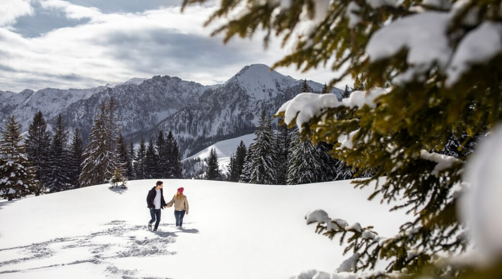 Winter couple (c) SalzburgerLand Tourismus Two people walk holding hands through snowy mountains surrounded by pine trees under a cloudy sky. (Enlarged view)
