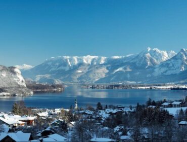 Wolfgangsee Winter St. Gilgen Snow-covered village by a lake with mountains in the background under a clear blue sky.
