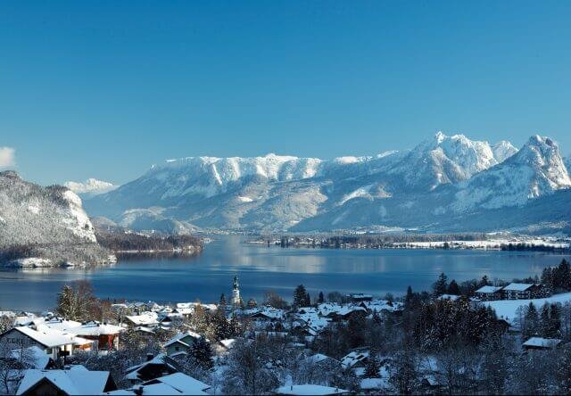 Snow-covered village by a lake with mountains in the background under a clear blue sky.