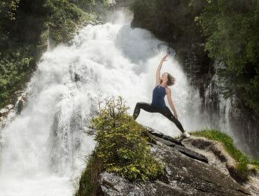 Woman doing a yoga pose on a rocky ledge by a rushing Gastein waterfall, surrounded by lush greenery.