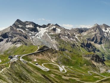 Winding mountain road with sharp bends, surrounded by green hills and snow-capped peaks under a clear sky.