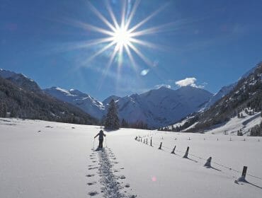 Person walks in snowy valley towards mountains under bright sun and clear blue sky, leaving footprints behind.