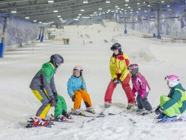 A ski instructor teaches four children inside an indoor ski slope, all wearing helmets and colourful gear.