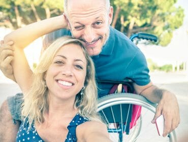 Smiling couple taking a selfie outdoors; one person is sitting in a wheelchair.