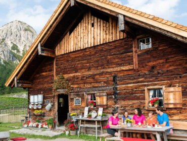 Tauernkarleitenalm (c) matthias-fritzenwallner(c)imatthias-fritzenwallner-12-1024×569 A family enjoys a meal at a wooden table outside their cabin, soaking in the charm of Alpine summer events.