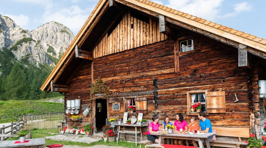 A family enjoys a meal at a wooden table outside their cabin, soaking in the charm of Alpine summer events.