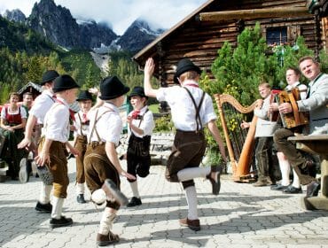 Boys in traditional Austrian attire dancing outdoors, with musicians and mountains in the background.