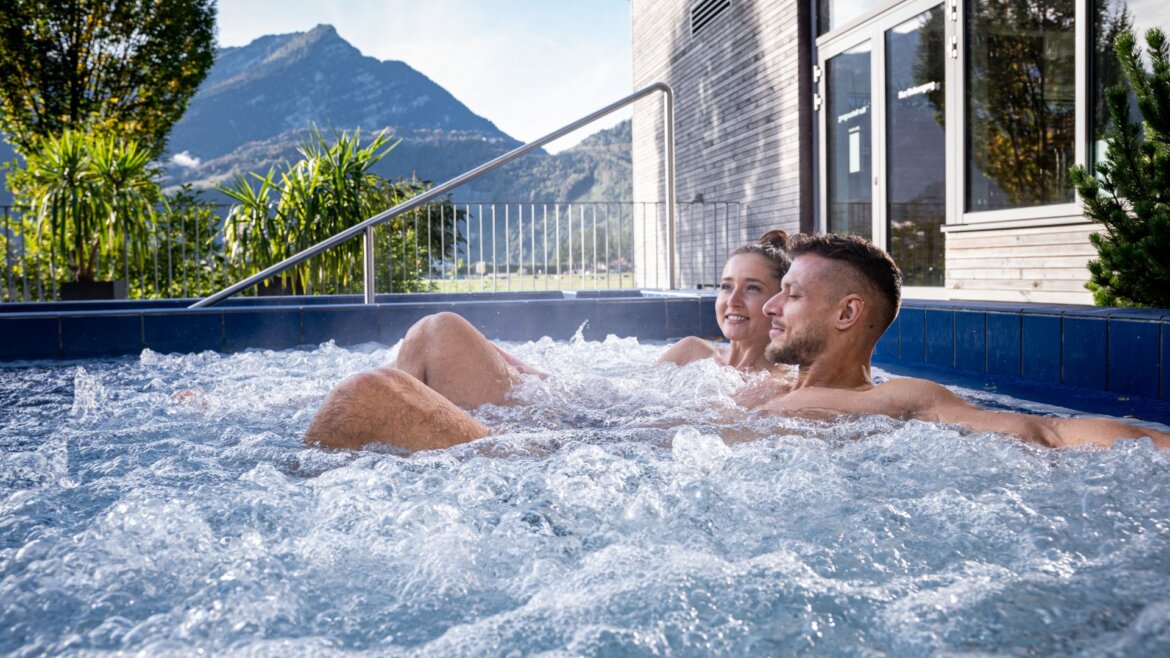 A couple relaxes in a bubbling outdoor hot tub with thermal water, surrounded by mountains and greenery.
