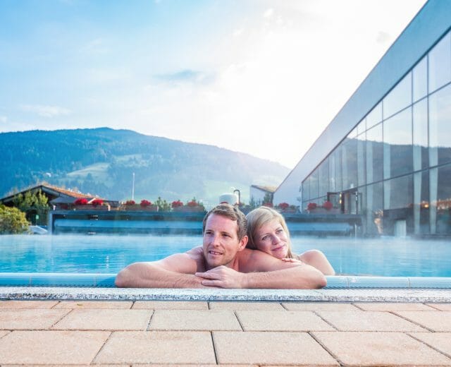 A couple relaxes in a thermal water pool with mountains and a modern spa building in the background.
