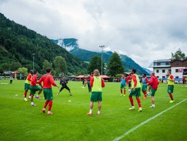 Football players in red and green kits warm up on a grassy pitch with mountains in the background.