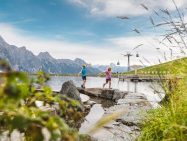 Two children balance on rocks by a lake, with mountains and cable cars in the background.