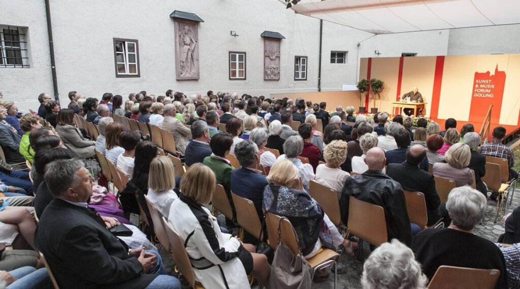A large seated audience watches a speaker at an indoor event with red and white decor.