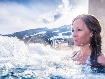 Woman enjoying a wellness retreat in an outdoor swimming pool with snowy mountains in the background on a sunny day.