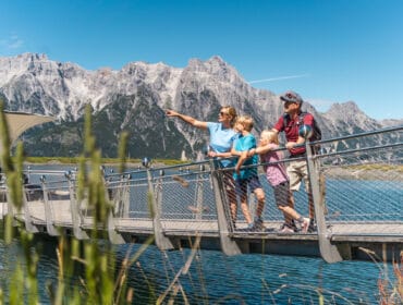 A family of four stands on a bridge by a lake, with mountains in the background under a clear sky.