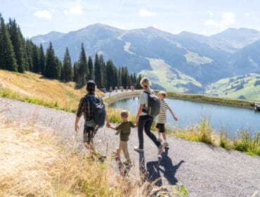 highres_saalbach_family_kodok_2023_DSC06876_47(c)karinpasterer.com A family of four hikes on a mountain path beside a lake with scenic hills in the background.