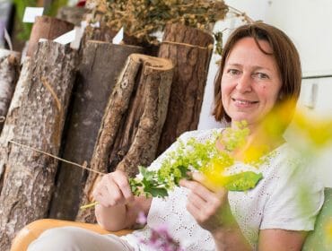 Woman sitting and smiling, holding green plants, evokes Traditional European Healing with logs and dried herbs behind her.