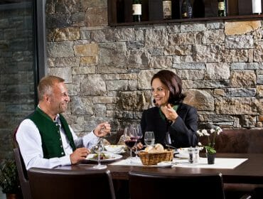 Two people dining at Via Culinaria, smiling and enjoying a meal with wine on the culinary pathway for gourmets.