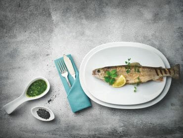 A must-try for Fish Fans: whole cooked fish with lemon, herbs, pesto, and black salt on a grey plate.