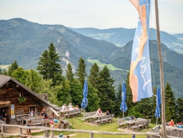 People enjoying Alpine summer events at outdoor tables by a wooden cabin, hills and valleys in the background.