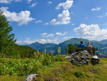 Two people sit on a bench in Obertauern, gazing at green mountains beneath a blue sky with scattered clouds.