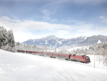 A red train ÖBB travels along snowy tracks through an Österreich landscape with mountains in the background.