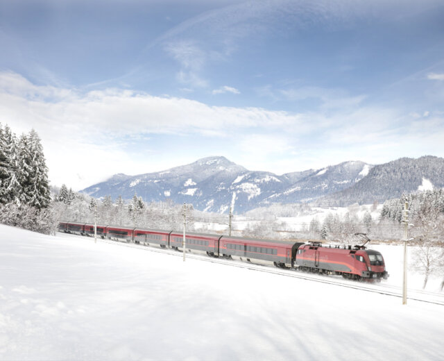 train winter (c) oebb A red train ÖBB travels along snowy tracks through an Österreich landscape with mountains in the background.