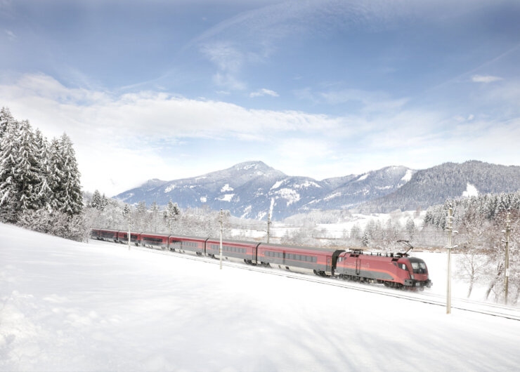 A red train ÖBB travels along snowy tracks through an Österreich landscape with mountains in the background.