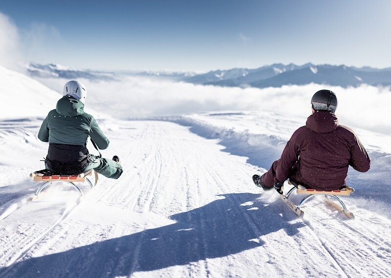 Two people sledging downhill on a snowy mountain with distant peaks and a clear sky in the background.